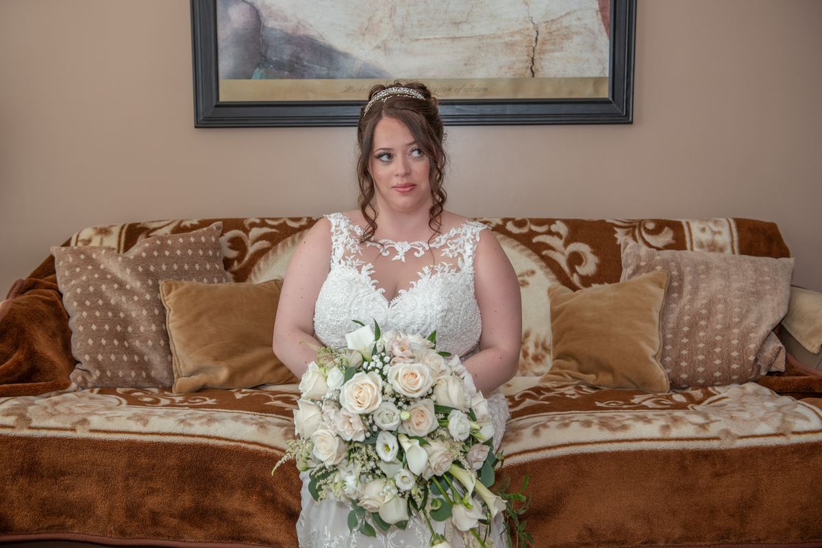 A bride in a white lace gown and floral bouquet sits thoughtfully on a brown velvet sofa, with a framed painting on the wall.