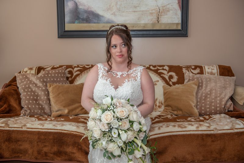 A bride in a white lace gown and floral bouquet sits thoughtfully on a brown velvet sofa, with a framed painting on the wall.