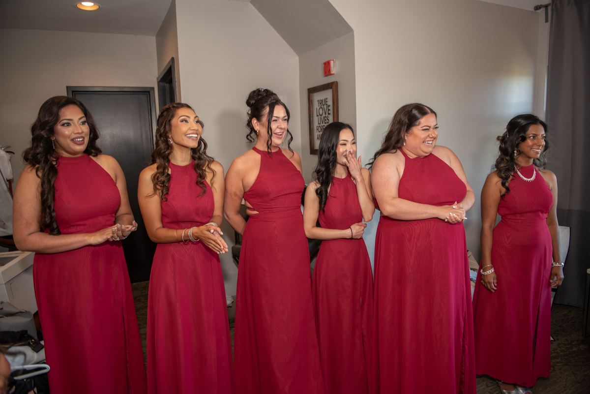 Bridesmaids in red gowns stand smiling and emotional, their expressions ranging from laughter to joyful tears.