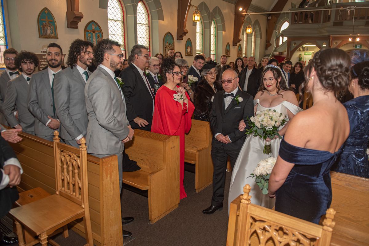 A bride in a white off-shoulder gown walks down the aisle, escorted by an older gentleman in a tuxedo.