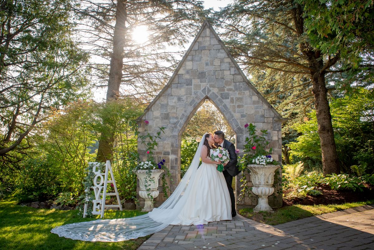A bride and groom kiss beneath a stone arch in a sunlit garden, surrounded by decorative urns and a LOVE sign.
