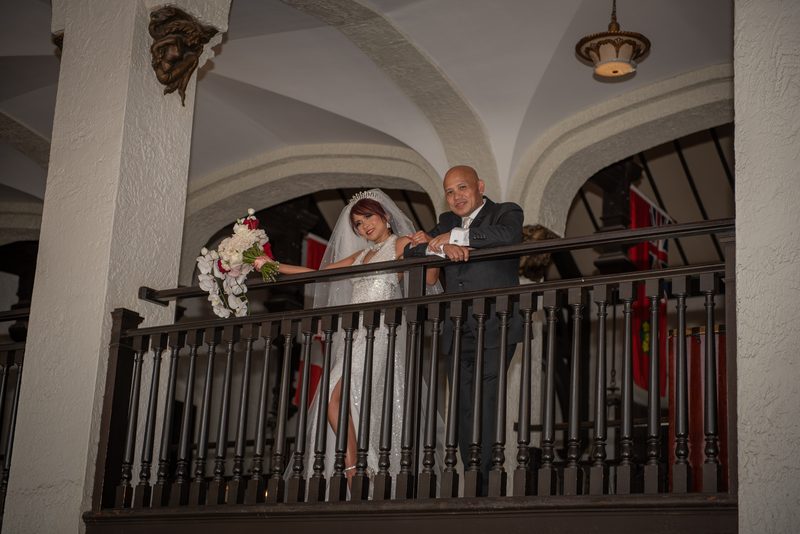 A bride in a white gown and a groom in a dark suit smile on an ornate balcony with arched ceilings and decorative columns.