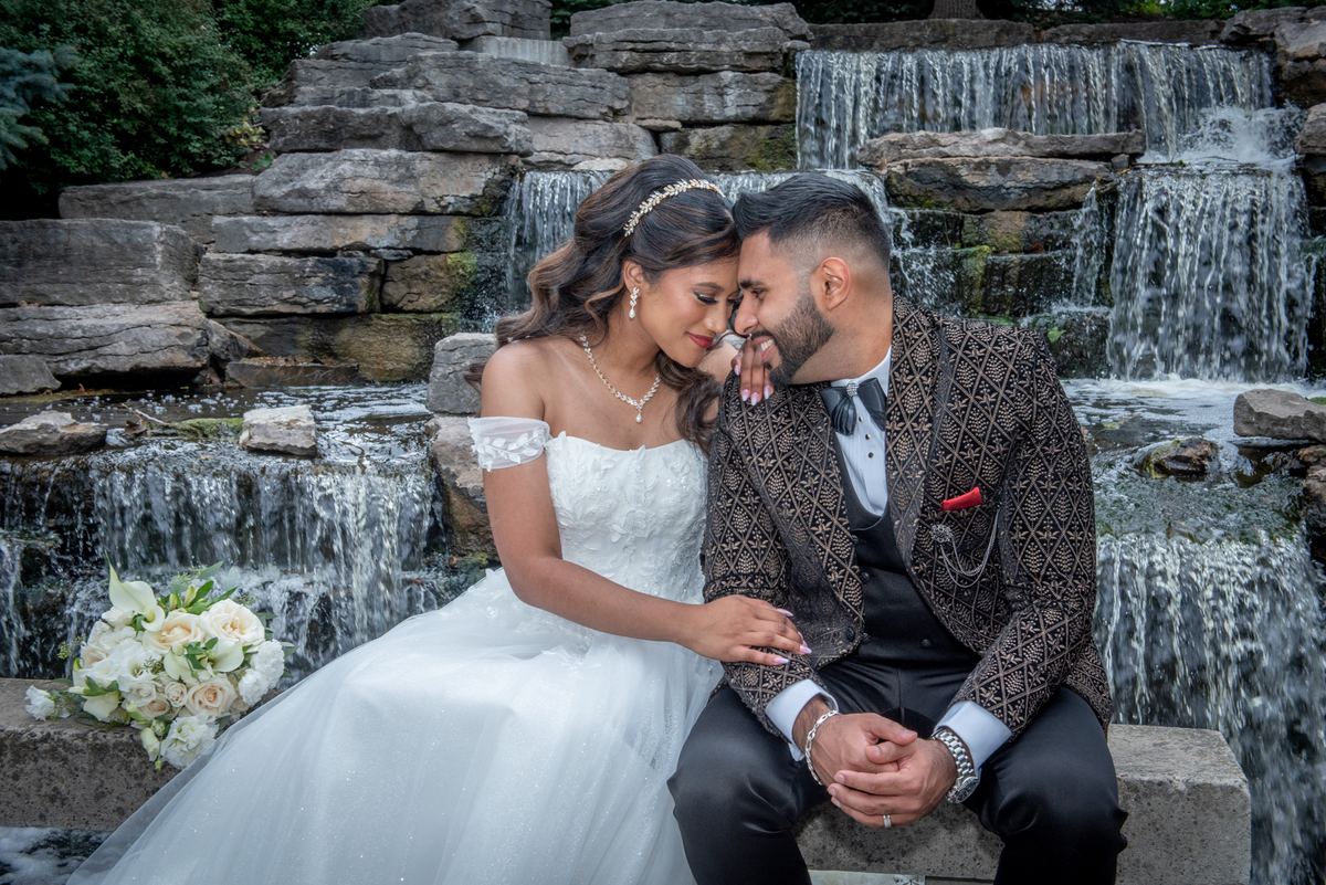 A newlywed couple sits by a waterfall, foreheads touching and hands intertwined, illuminated by soft light.
