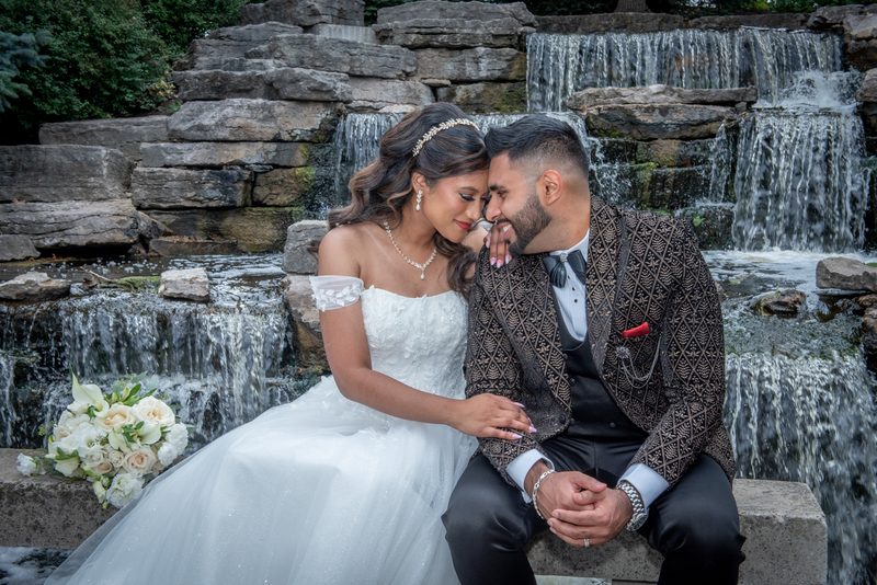 A newlywed couple sits by a waterfall, foreheads touching and hands intertwined, illuminated by soft light.
