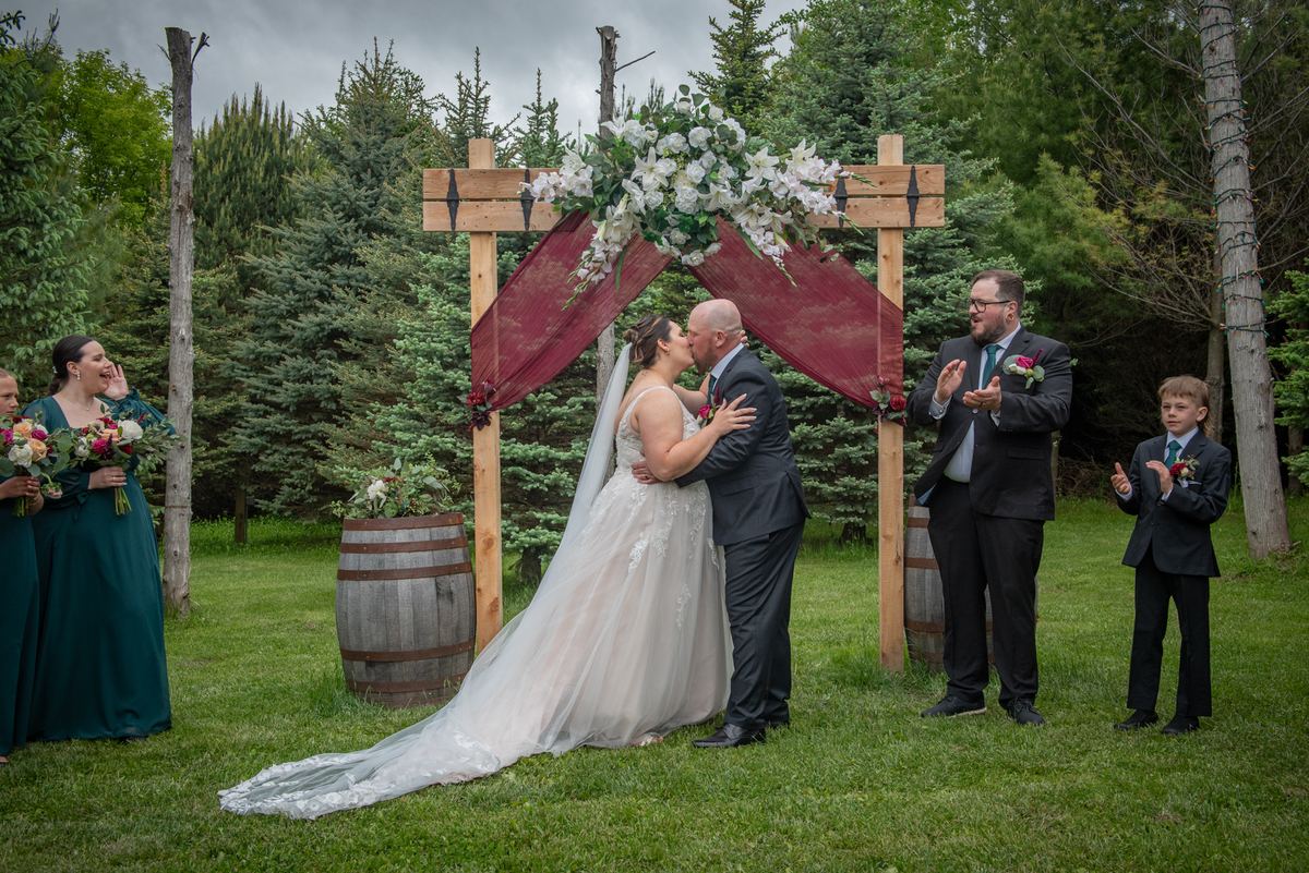 Bride and groom kiss under wooden arch with white flowers and burgundy fabric as guests celebrate outdoors.