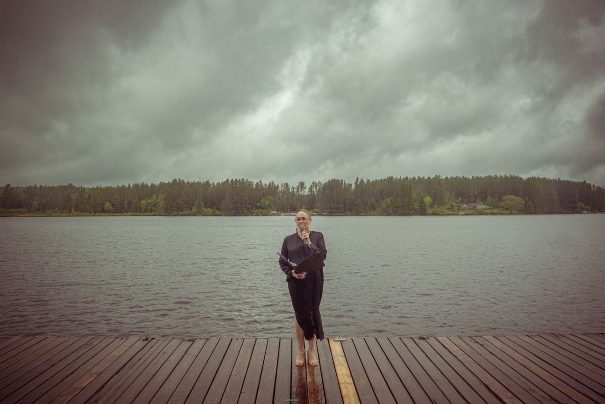 A woman stands alone on a wooden dock by a calm lake, holding a microphone and folder under an overcast sky.