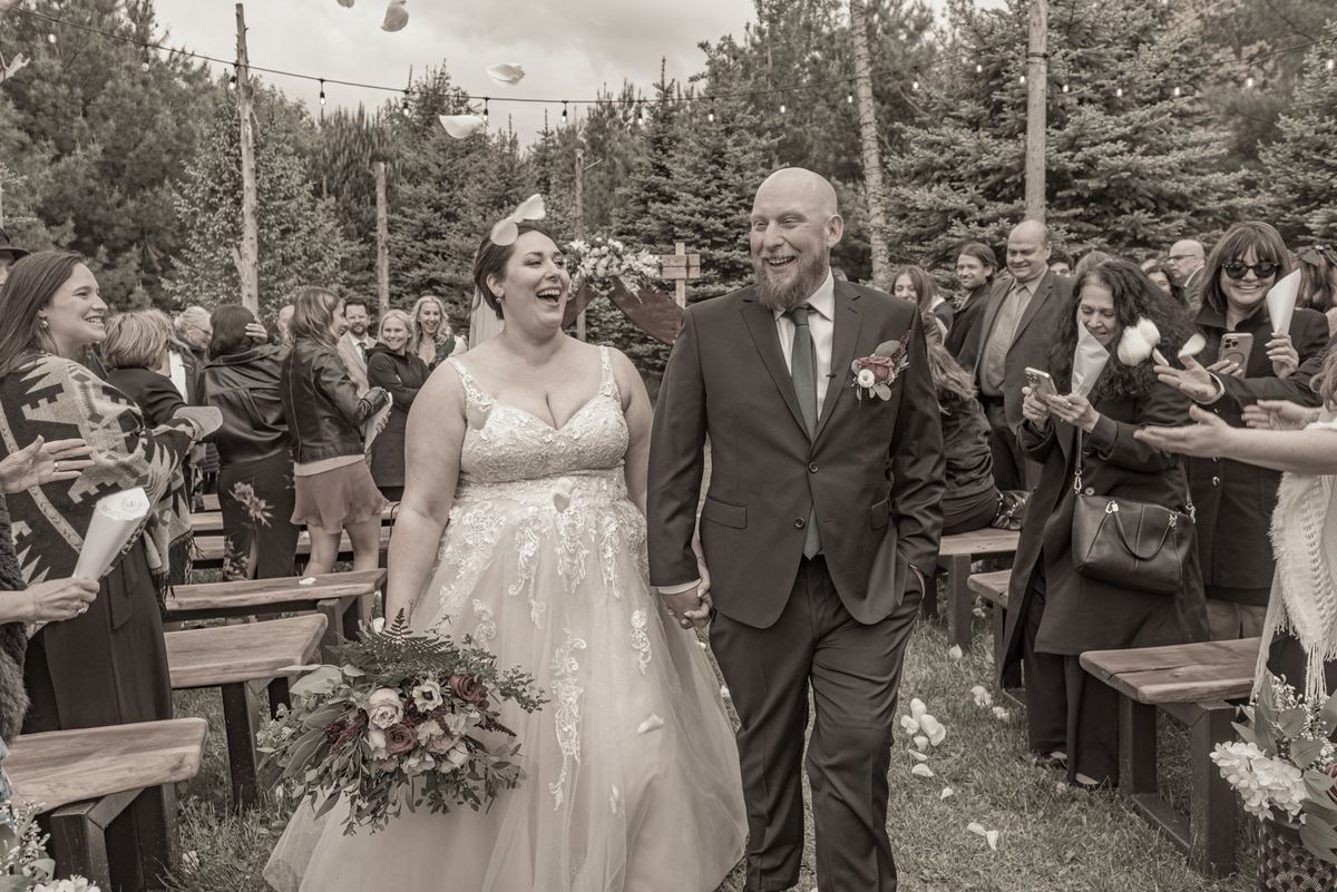 A smiling bride and groom walk down the aisle in a forest, surrounded by guests tossing petals on a gray day.