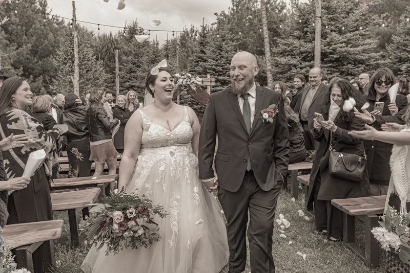 A smiling bride and groom walk down the aisle in a forest, surrounded by guests tossing petals on a gray day.