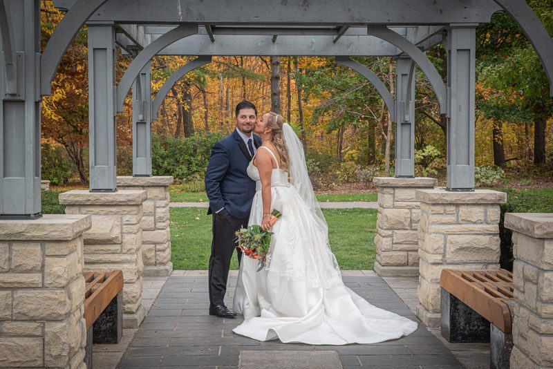 A bride in formal attire kisses her groom beneath an arbor, surrounded by autumn trees and soft daylight.