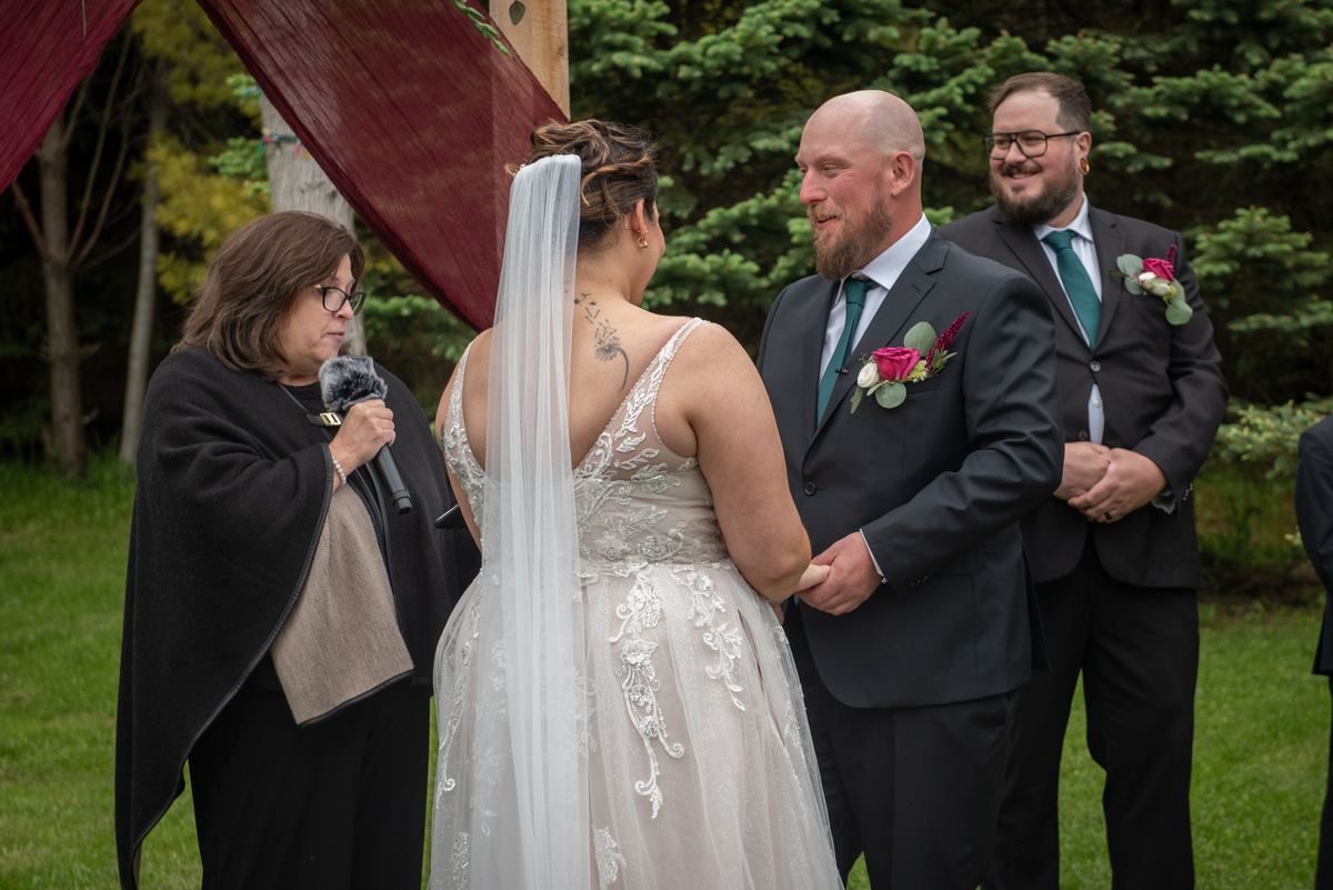 Bride in lace dress and veil, groom in dark suit, exchange vows under draped canopy with officiant and groomsman present.