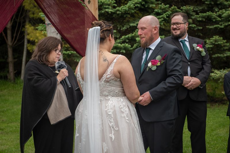 Bride in lace dress and veil, groom in dark suit, exchange vows under draped canopy with officiant and groomsman present.