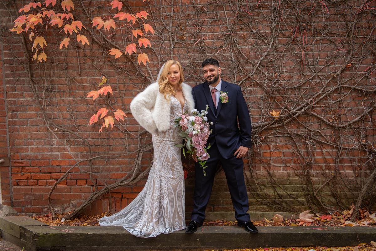 Bride in lace gown and fur stole with groom in dark suit stand by rustic brick wall covered in autumn vines.