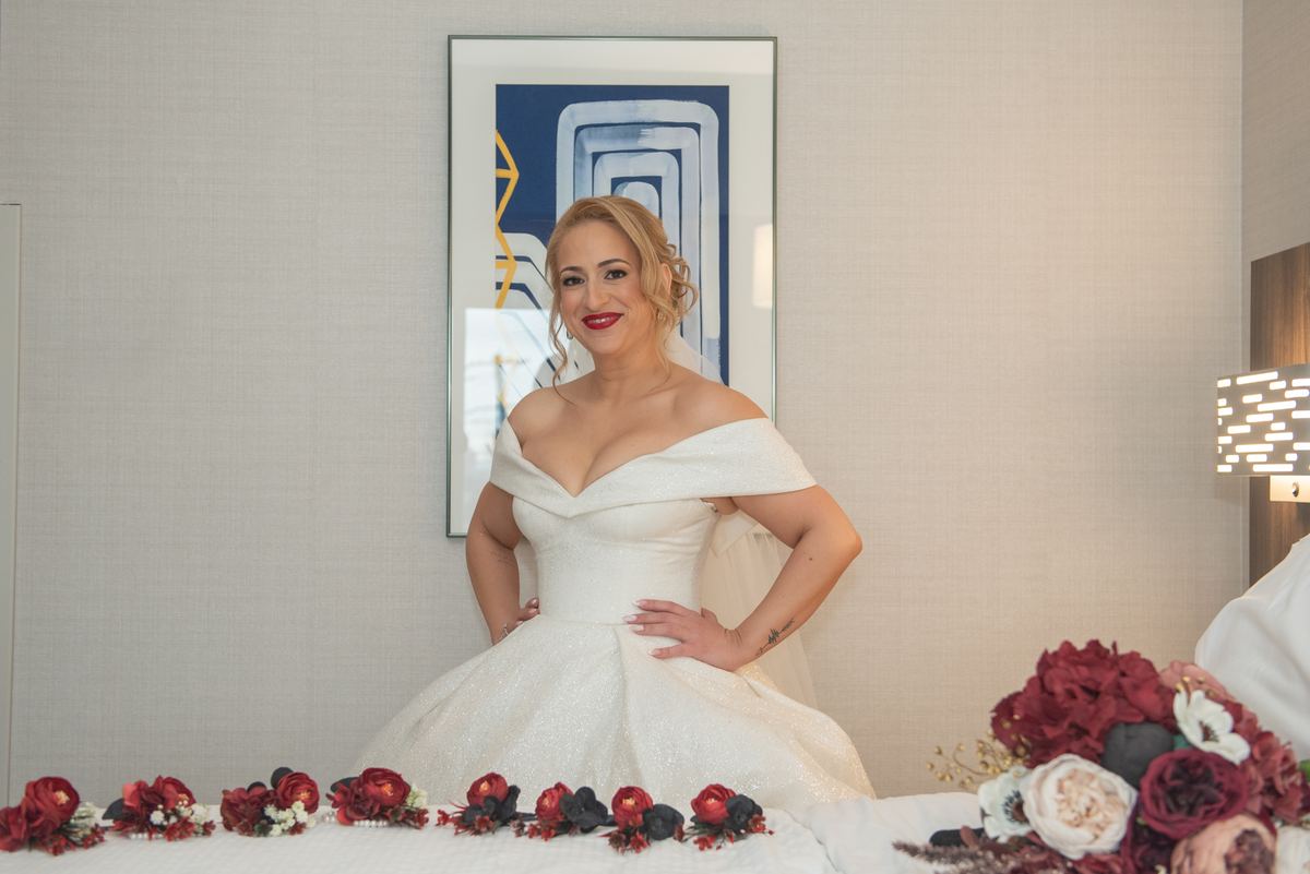 A bride in an off-the-shoulder gown stands confidently by a bed covered with red flowers and modern art on the wall.