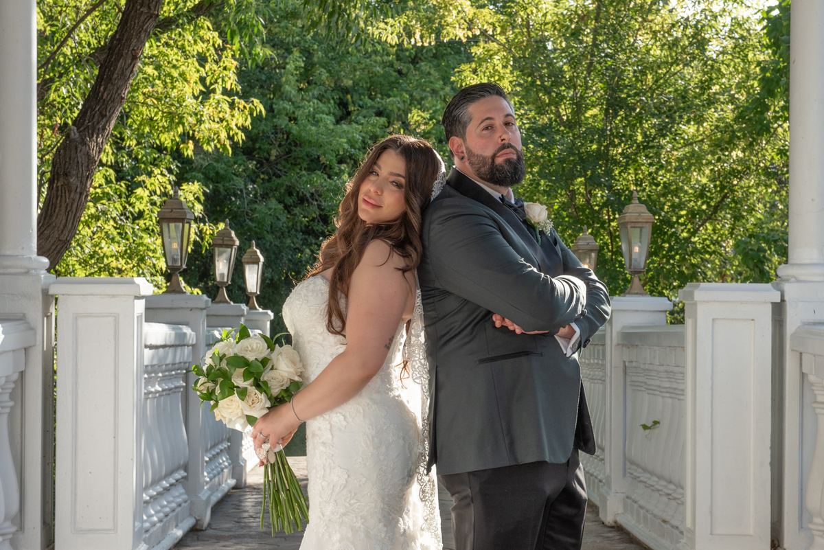 Bride in lace gown and groom in grey suit stand back-to-back on porch, smiling at camera and looking aside.
