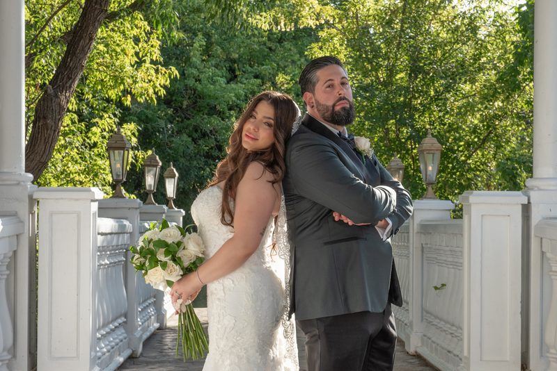 Bride in lace gown and groom in grey suit stand back-to-back on porch, smiling at camera and looking aside.