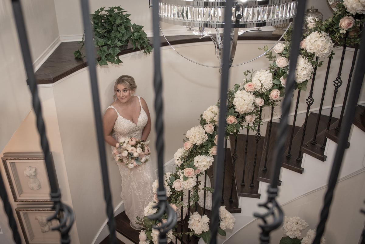 A bride in a lace gown stands on a grand staircase, framed by iron balusters and floral arrangements.