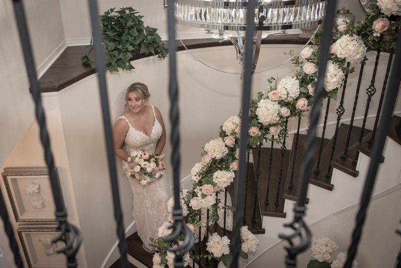 A bride in a lace gown stands on a grand staircase, framed by iron balusters and floral arrangements.