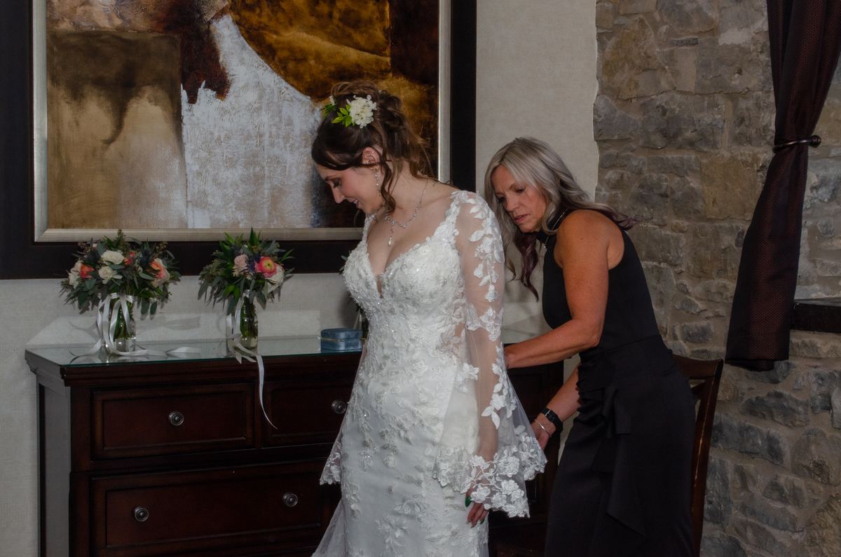 A bride in an ornate lace gown is being helped by a woman who adjusts the back of her dress in a rustic room.
