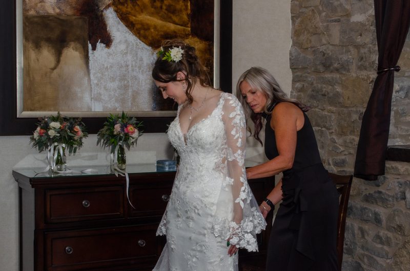 A bride in an ornate lace gown is being helped by a woman who adjusts the back of her dress in a rustic room.