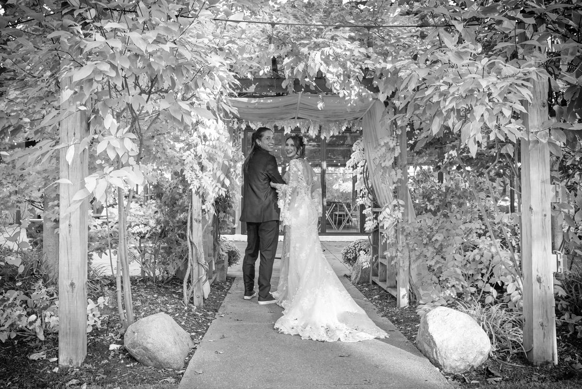A bride and groom smile at each other under a leafy, vine-covered arbor in an outdoor setting with diffused light.