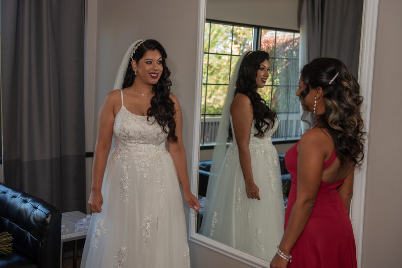 A bride in a white gown and veil smiles at her reflection next to a woman in red, bathed in soft natural light.