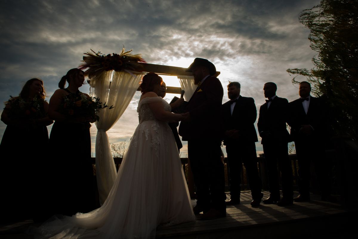 A bride and groom stand under a decorated arch during a ceremony with guests in formal attire around them.