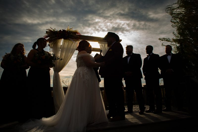 A bride and groom stand under a decorated arch during a ceremony with guests in formal attire around them.