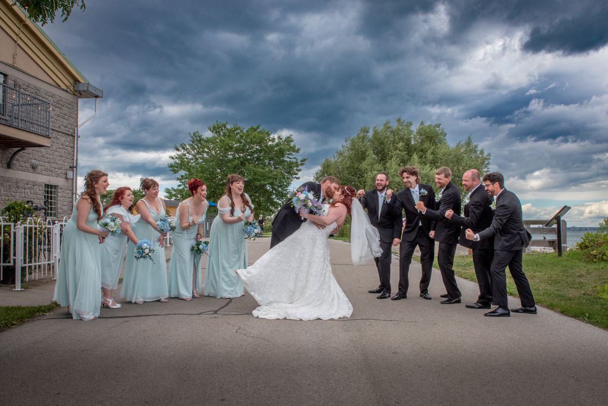 A bride and groom kiss passionately with the wedding party smiling in the background under an overcast sky near water.