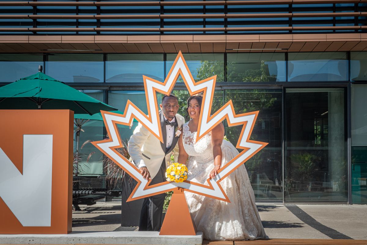 A bride in a lace gown and groom in a white tuxedo pose behind a large maple leaf cutout on a sunny patio.