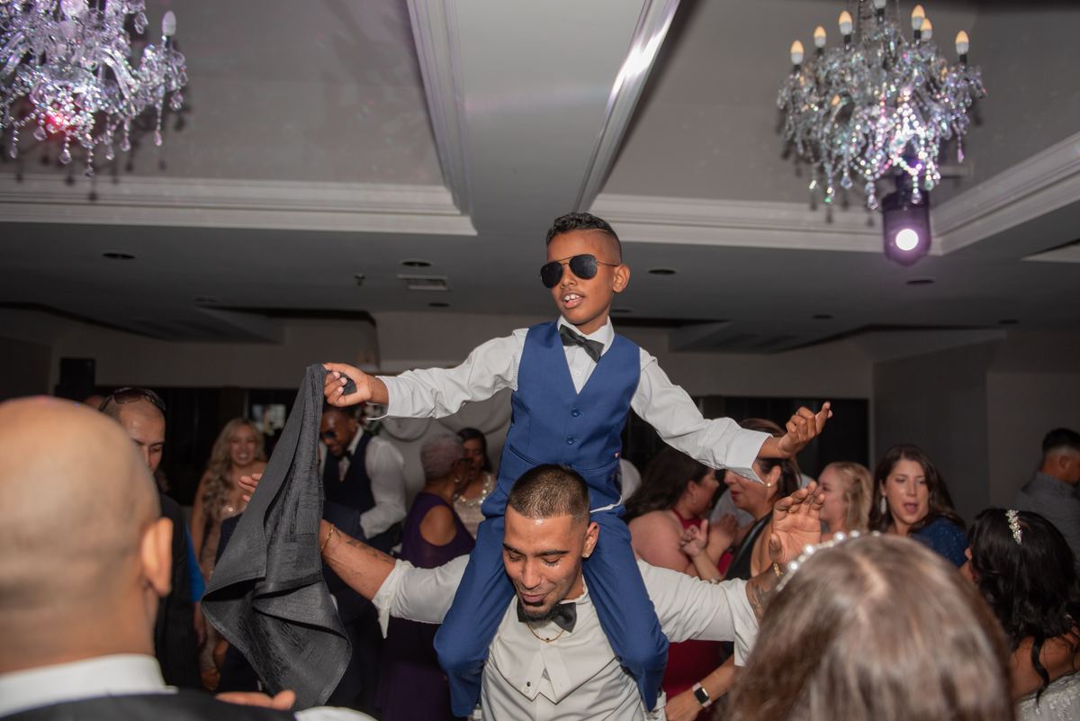 A young boy in a blue vest and sunglasses dances on an adult's shoulders at a lively wedding reception.