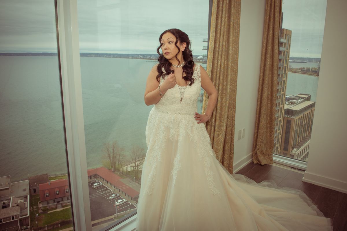 A bride in a lace gown stands by a window, adjusting her dress as she looks out at an overcast cityscape.