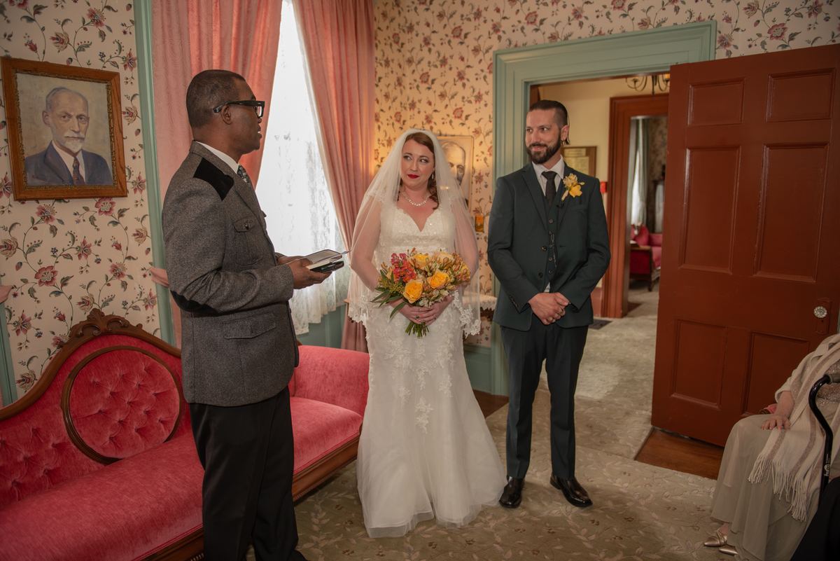 A bride and groom stand facing each other during a ceremony in a vintage room with floral wallpaper.