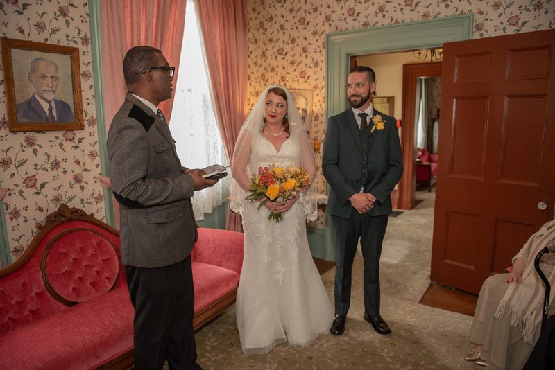 A bride and groom stand facing each other during a ceremony in a vintage room with floral wallpaper.
