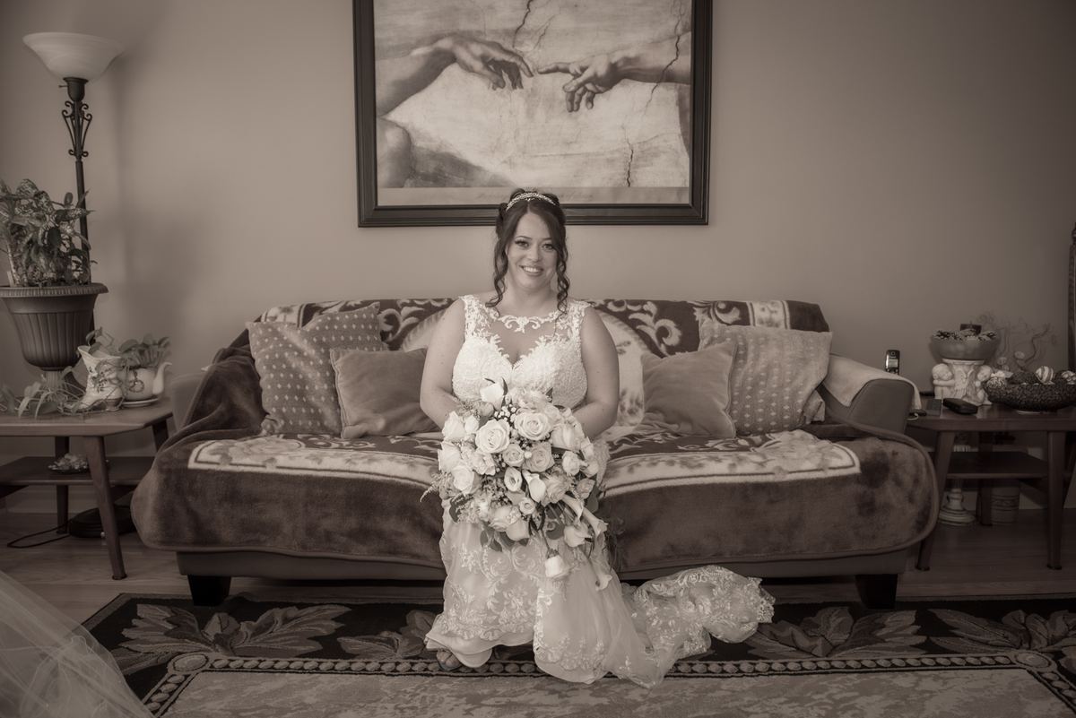 A smiling bride in a wedding dress kneels on a patterned rug in a cozy living room with a bouquet of flowers.