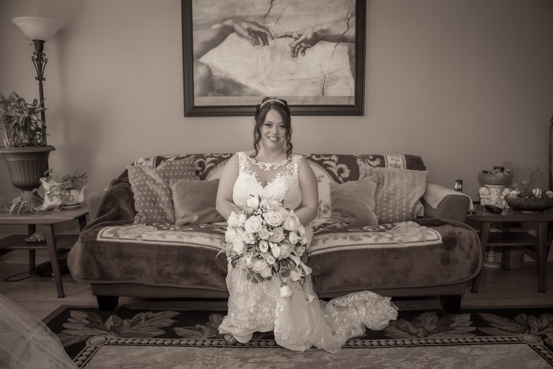 A smiling bride in a wedding dress kneels on a patterned rug in a cozy living room with a bouquet of flowers.