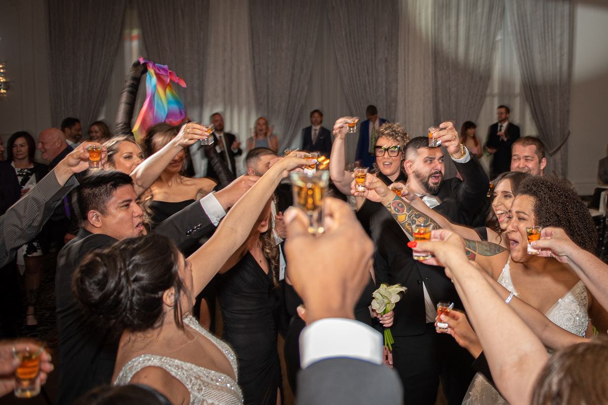 Guests toast the bride and groom in formal attire at a joyful celebration, with a colorful rainbow flag waving nearby.
