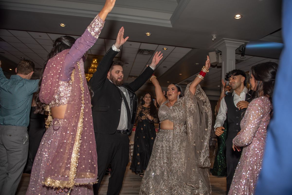 Guests in elegant attire dance joyfully with raised arms under warm ambient lights at a cultural celebration.