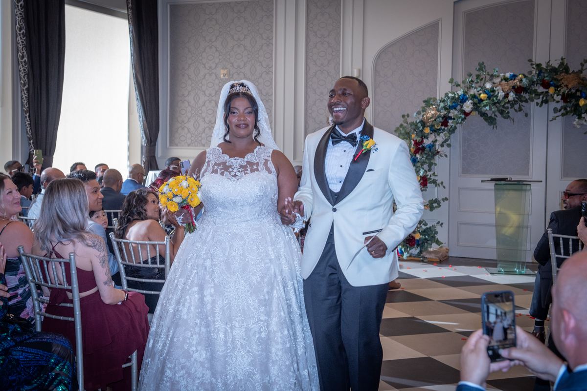 A smiling bride in a lace gown and veil walks hand-in-hand with her groom in a white tuxedo down the aisle towards guests.