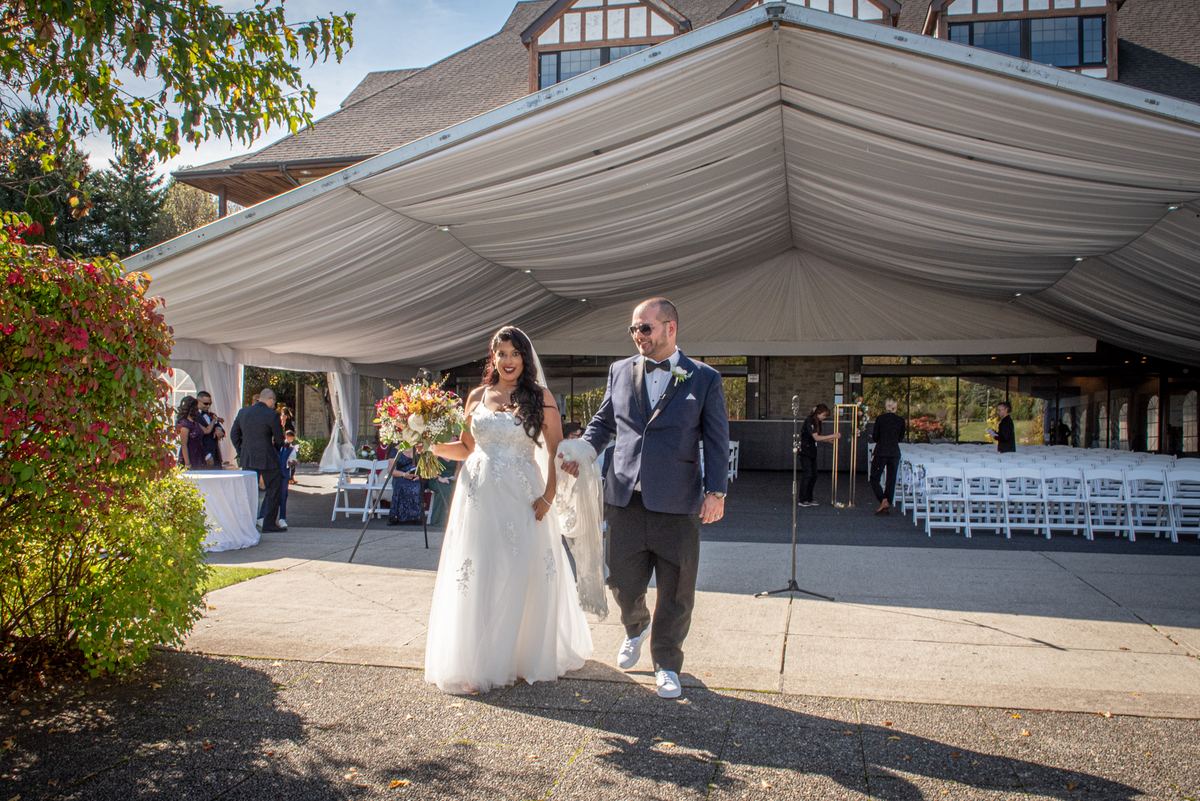 A bride in a white gown walks confidently down the aisle toward a draped tent, escorted by a man in a dark suit.
