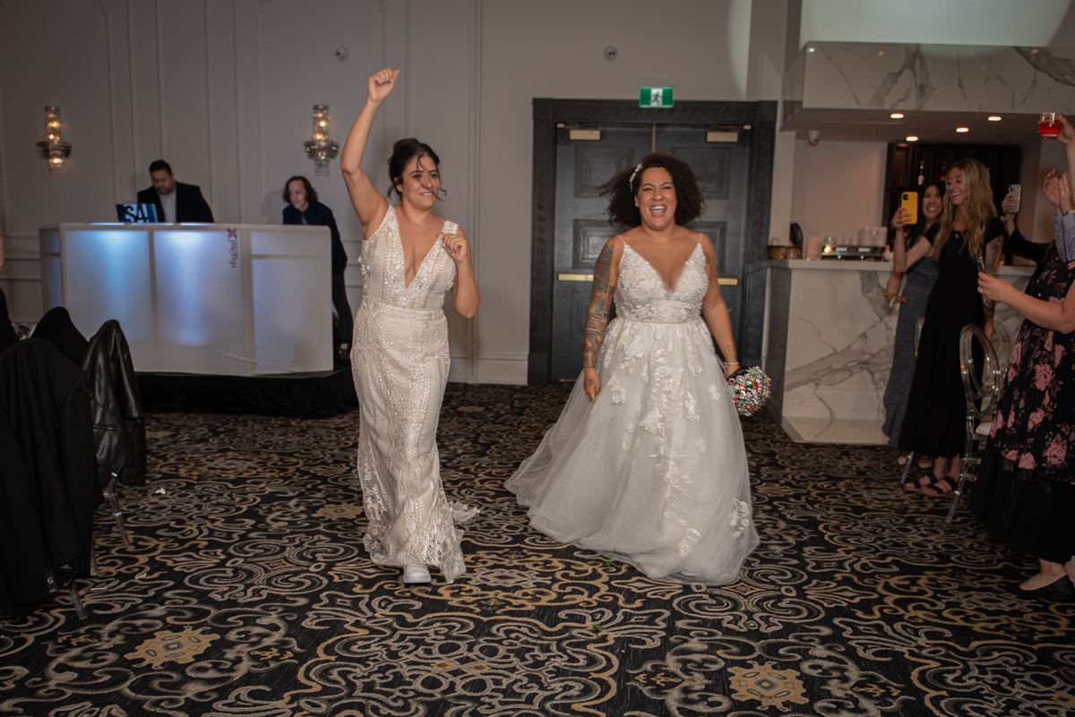 Two brides smiling and raising a fist, walking down a carpeted aisle toward the camera in warm indoor lighting.
