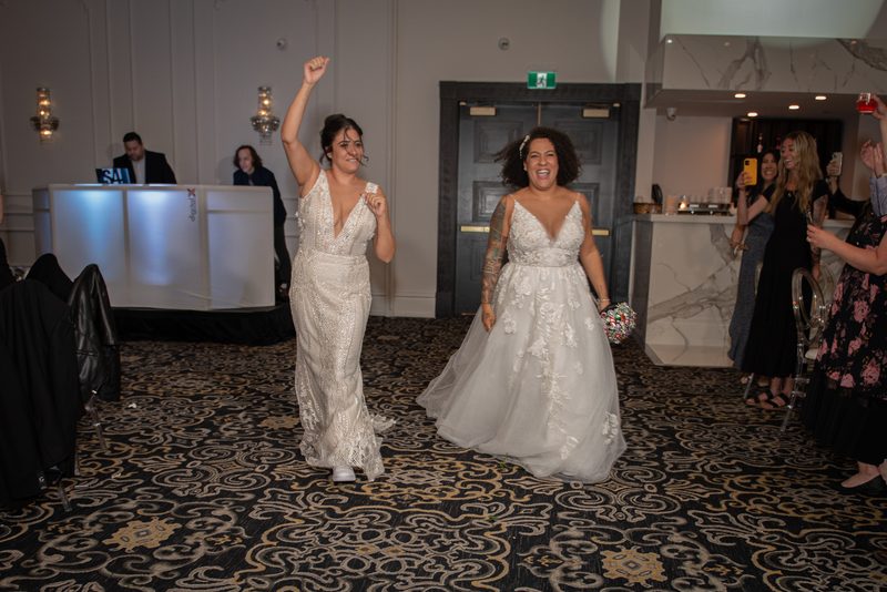 Two brides smiling and raising a fist, walking down a carpeted aisle toward the camera in warm indoor lighting.