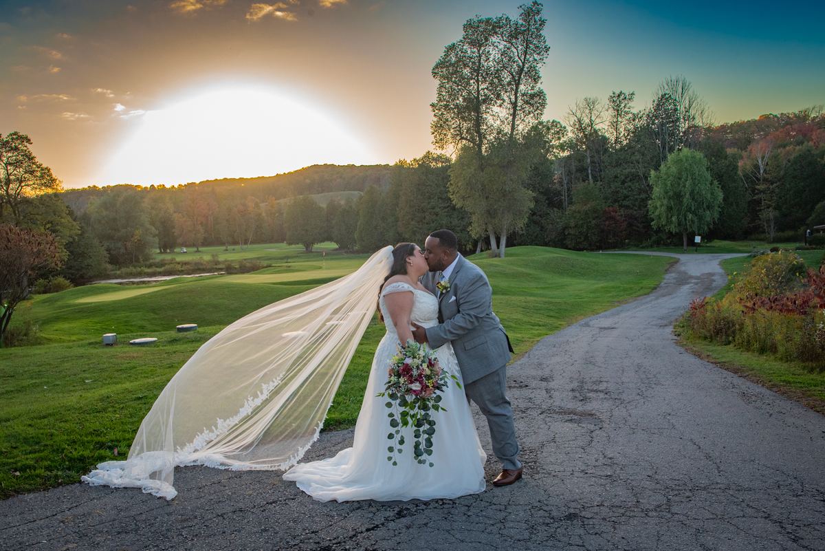 A bride and groom kiss beside a golf course path as the sun sets, their long shadows stretching across the ground.