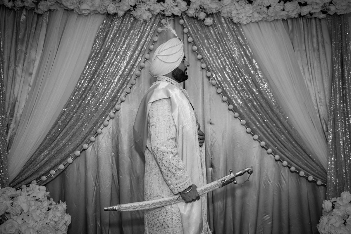 A Sikh groom in ceremonial attire holds a kirpan against a backdrop of flowers and soft lighting, exuding dignity.