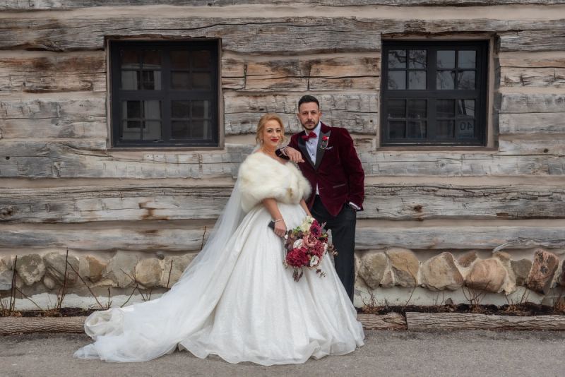 A bride in a white gown and fur stole poses with her groom in a burgundy tuxedo outside a rustic cabin.