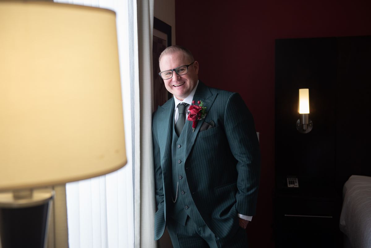 A groom in a pinstripe suit and red boutonniere leans against a doorway, smiling warmly in soft light.