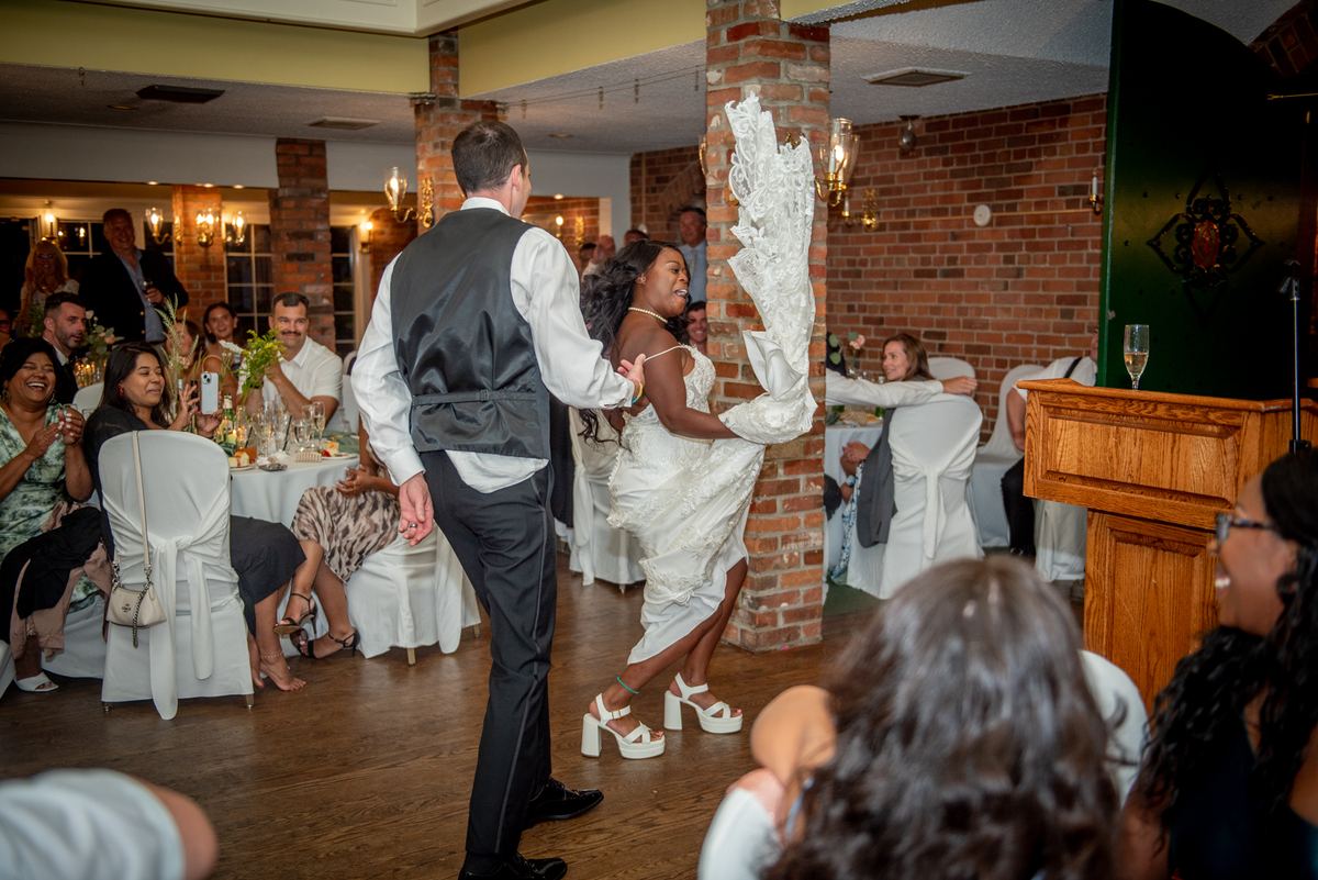 A bride and groom dance joyfully, she lifting her veil as he holds her hand, guests watching and laughing.