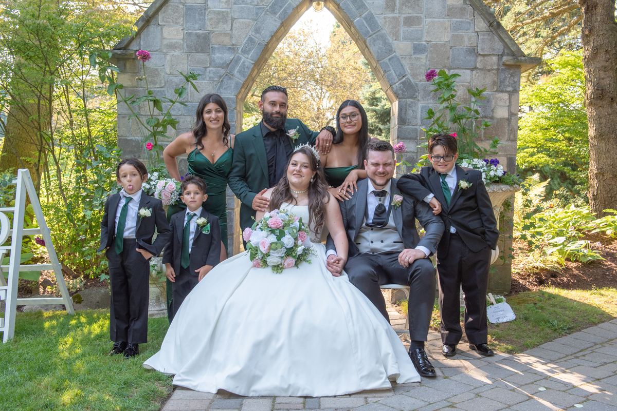A wedding party poses under a stone archway in a garden, surrounded by floral arrangements and family members.