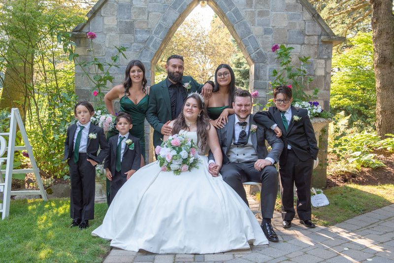 A wedding party poses under a stone archway in a garden, surrounded by floral arrangements and family members.
