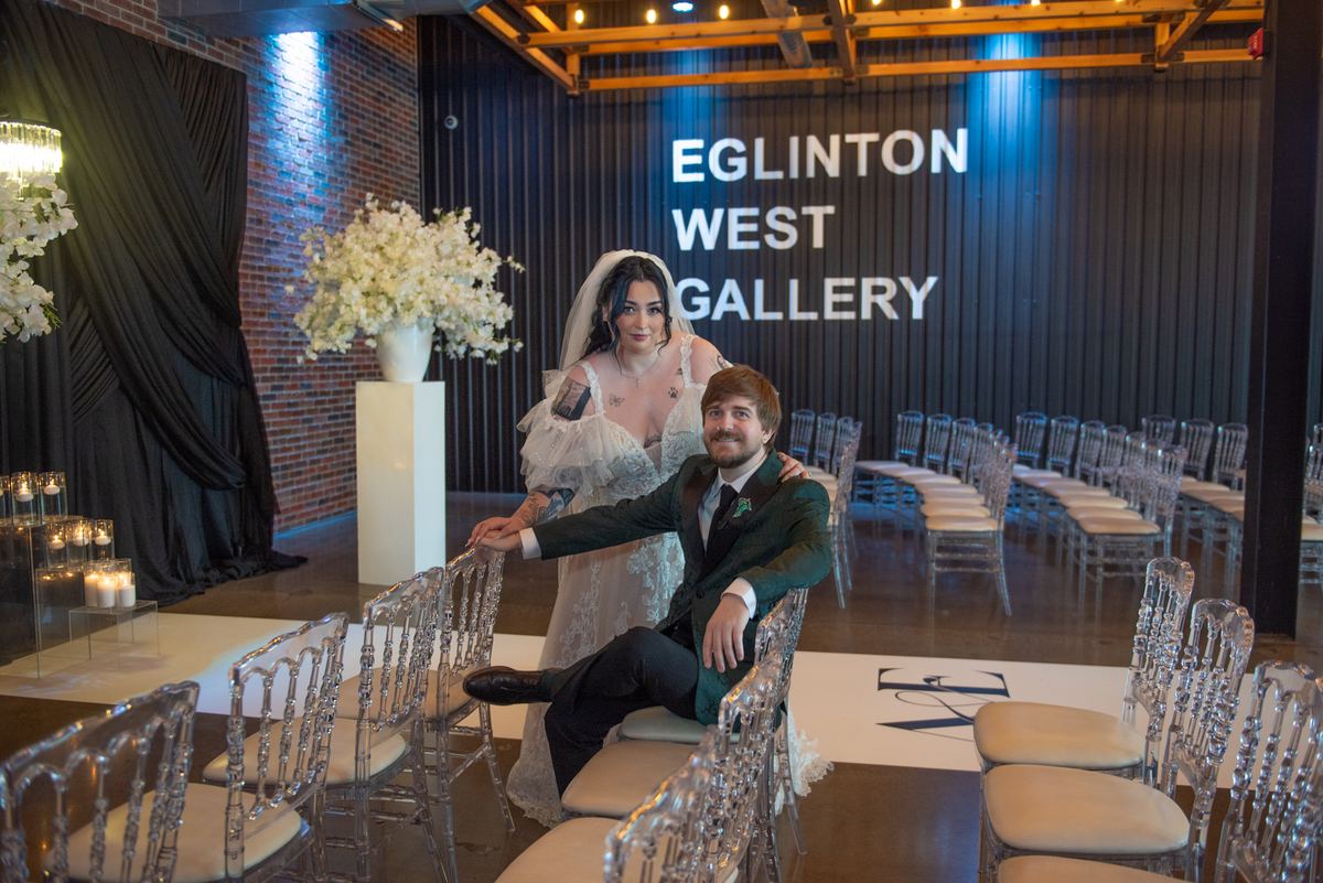 A bride in a lace gown stands behind her seated groom with outstretched arm, surrounded by elegant decor.