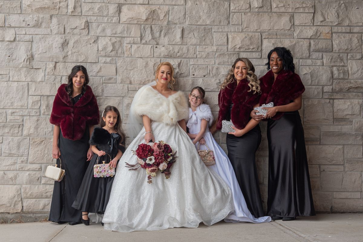A bridal party poses joyfully against a stone wall, the bride in white with a burgundy bouquet, bridesmaids in black gowns.